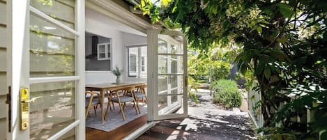 An open door leading to a covered outdoor area with a table and chairs, surrounded by greenery.