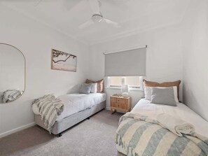 A bedroom with two beds, a ceiling fan, a window with blinds, a bedside table, and a wall-mounted mirror. - Newberry Cottage: Beach, Games & Alfresco Bliss (Dunsborough)