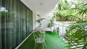 Un balcón con una pequeña mesa y una silla, rodeado de abundante vegetación y un muro de cristal.