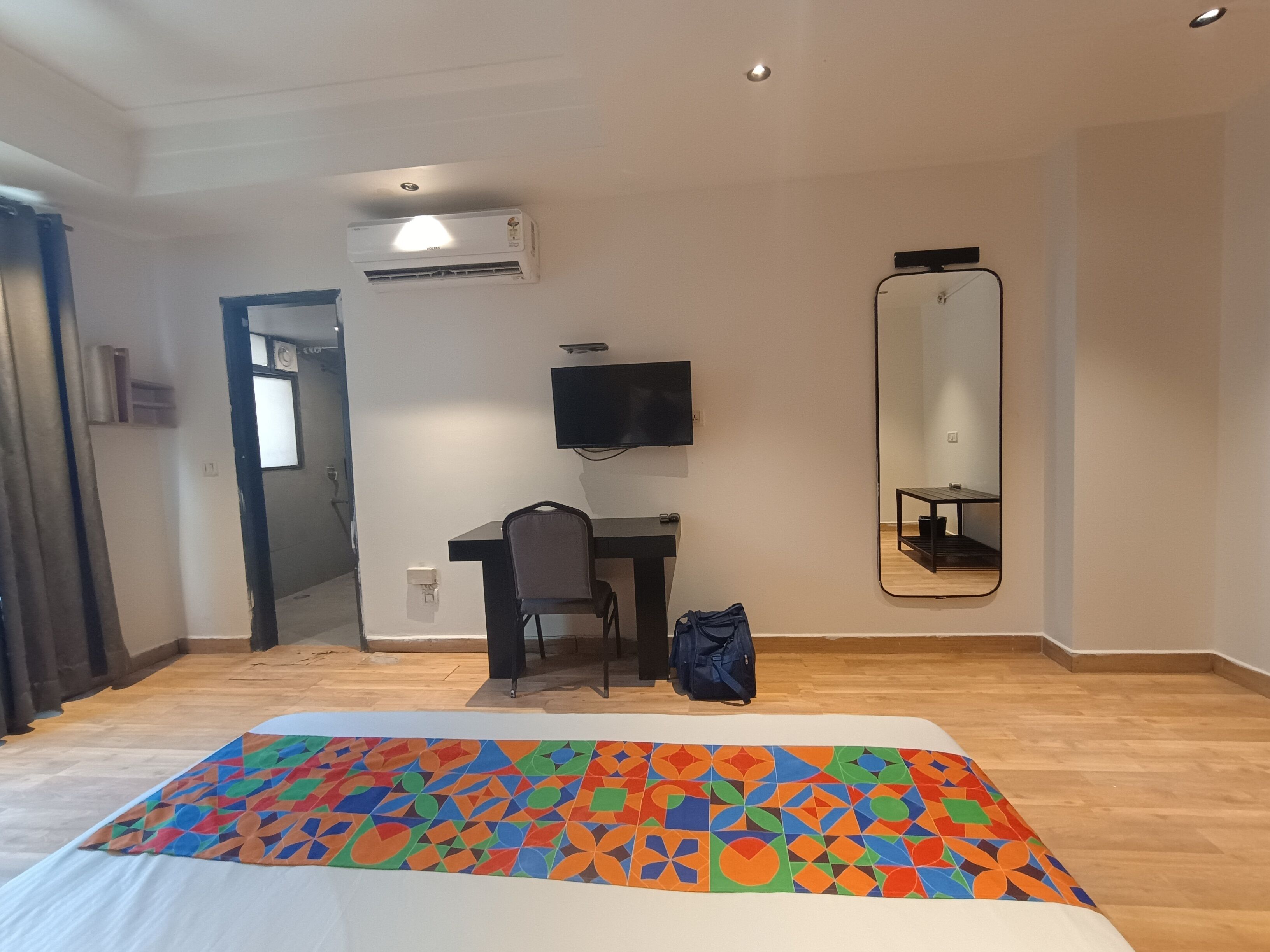 A hotel room with a bed, a desk, a chair, a TV, a mirror, and an air conditioning unit.