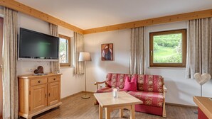 Un salon chaleureux avec un plancher en bois, un canapé rouge agrémenté de coussins à motifs floraux, un meuble TV en bois, une table avec une bougie et une fenêtre donnant sur un paysage verdoyant.