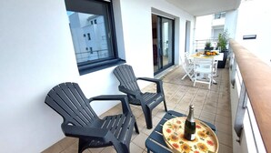 A balcony with two black chairs, a small table with a bottle and glass, and a dining area with a table and chairs.