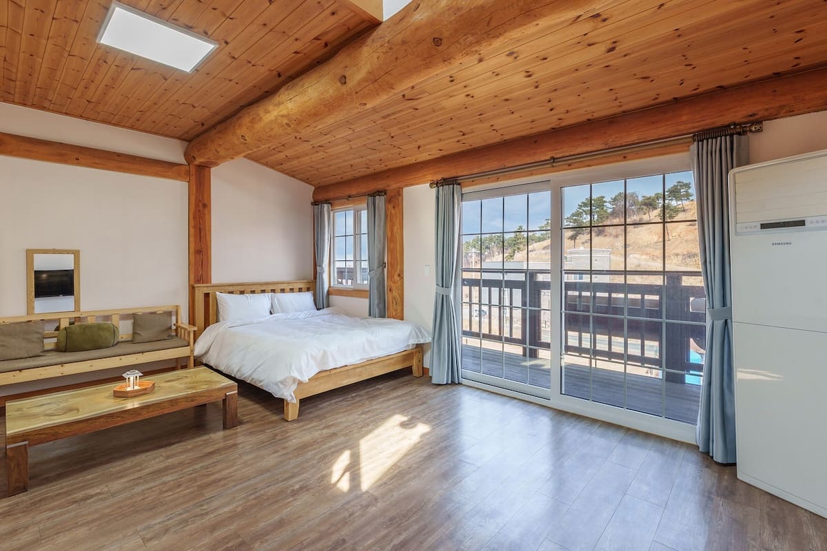 Un dormitorio con techo de madera, una cama, un banco, un espejo y un ventanal con vistas al exterior.