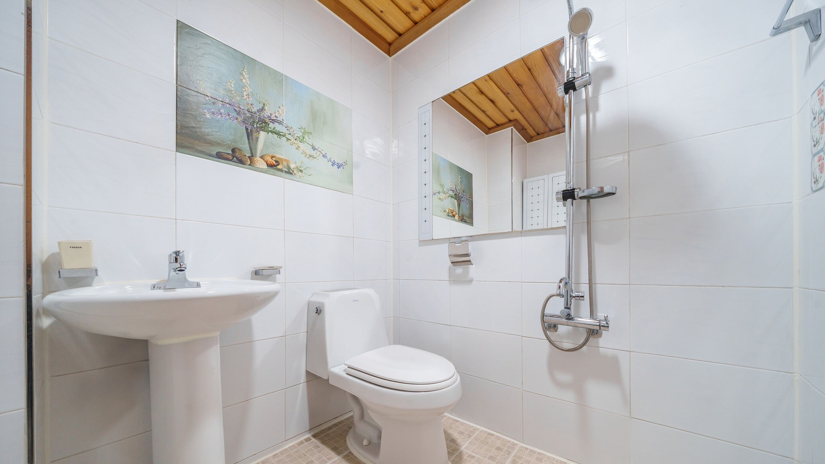 A bathroom with a white sink, toilet, and shower. There is a painting on the wall above the sink and a mirror above the shower.