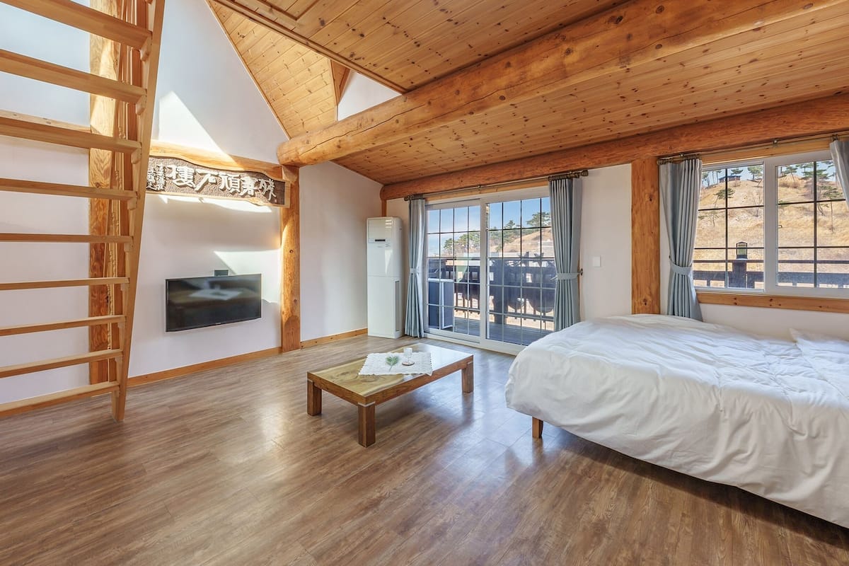 Una habitación con una cama, un televisor montado en la pared, una mesa de madera y un ventanal corredero que da a un espacio exterior.