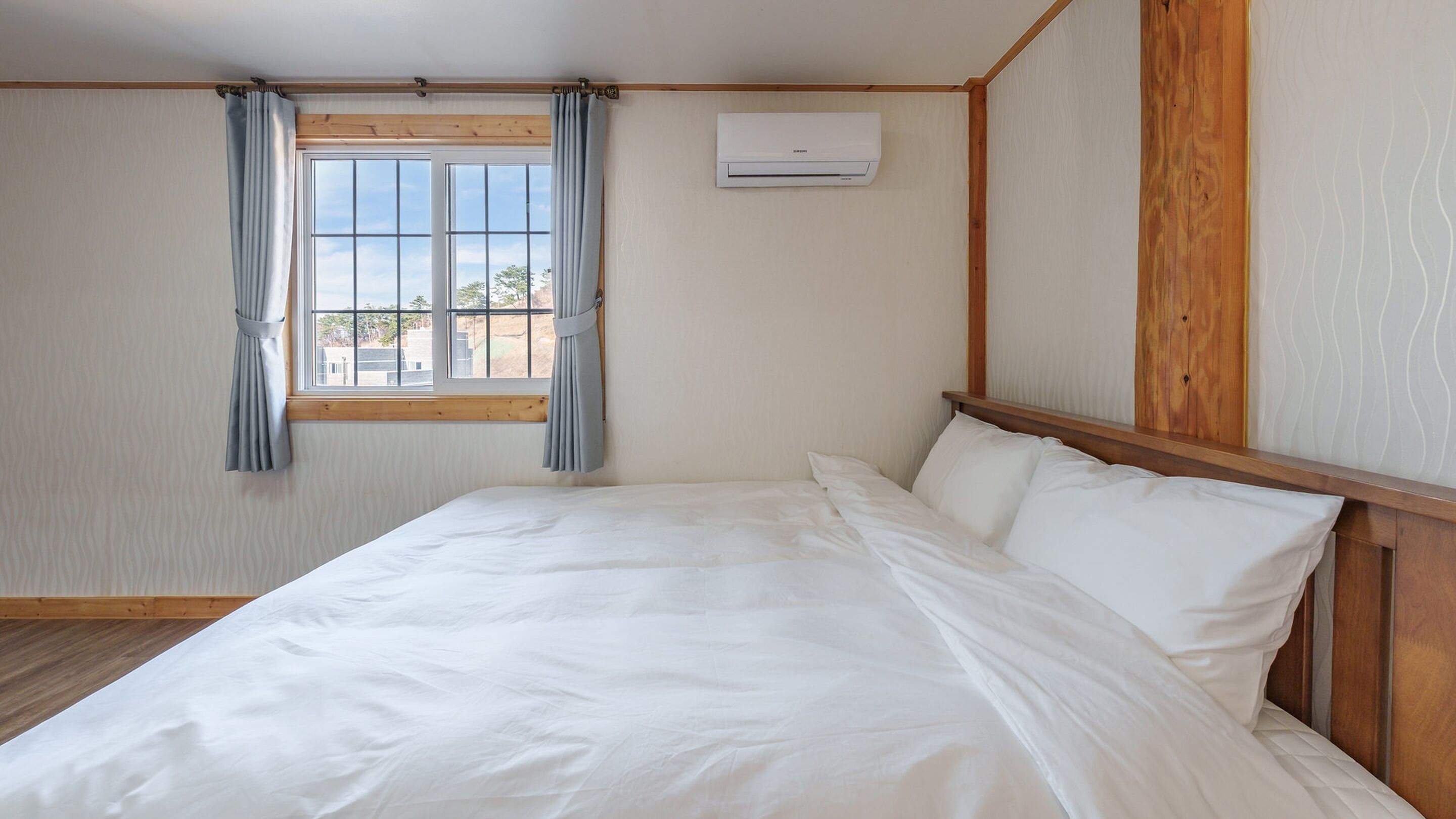 Un dormitorio con cabecera de madera, una cama individual con ropa de cama blanca, una ventana con cortinas y un equipo de aire acondicionado en la pared.