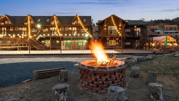 A rustic lodge with a large fire pit in the foreground, wooden buildings with multiple windows and a covered outdoor seating area in the background. - Daecheon Ocean Forest Pension (Boryeong)