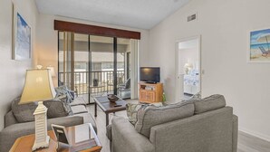 A living room with a sofa, a coffee table, a TV on a wooden cabinet, and a balcony with a view of the outside.