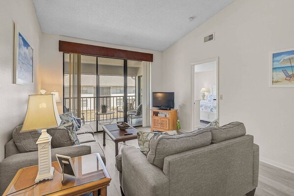 A living room with a sofa, a coffee table, a TV on a wooden cabinet, and a balcony with a view of the outside.