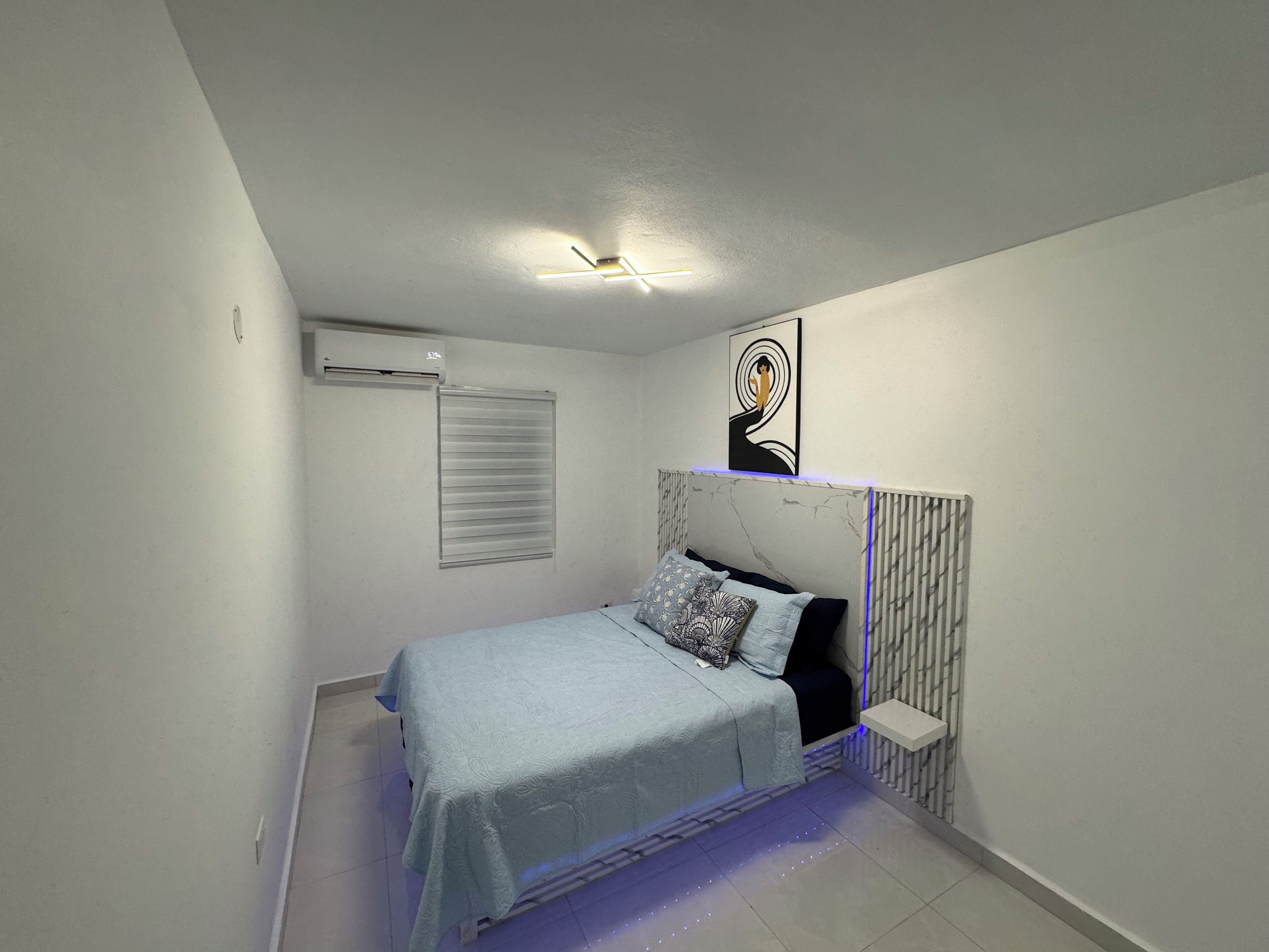A bedroom with a bed, a ceiling fan, an air conditioner, a wall-mounted shelf, and a decorative piece above the bed.