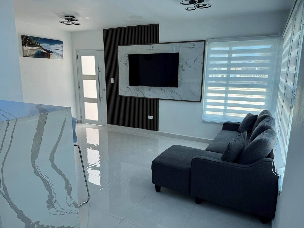 Una sala de estar moderna con una televisión de pantalla plana montada en una pared de mármol, un sofá gris y un amplio ventanal con persianas.