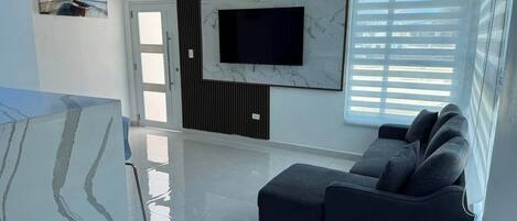 Una sala de estar moderna con una televisión de pantalla plana montada en una pared de mármol, un sofá gris y un amplio ventanal con persianas.