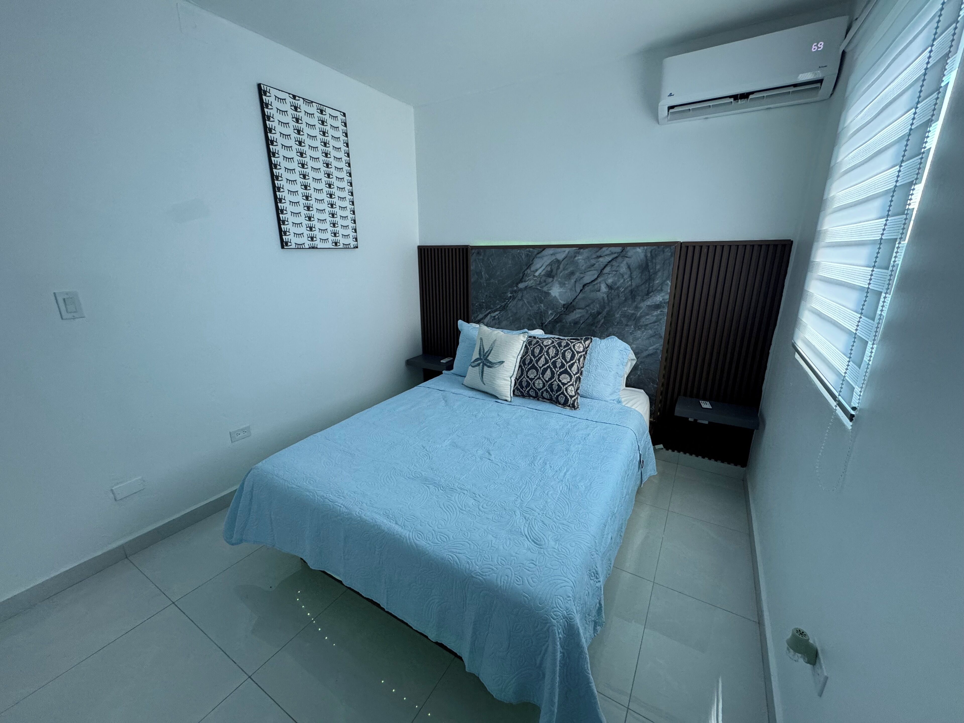 A modern bedroom with a bed, a headboard, an air conditioning unit, and a wall-mounted artwork.