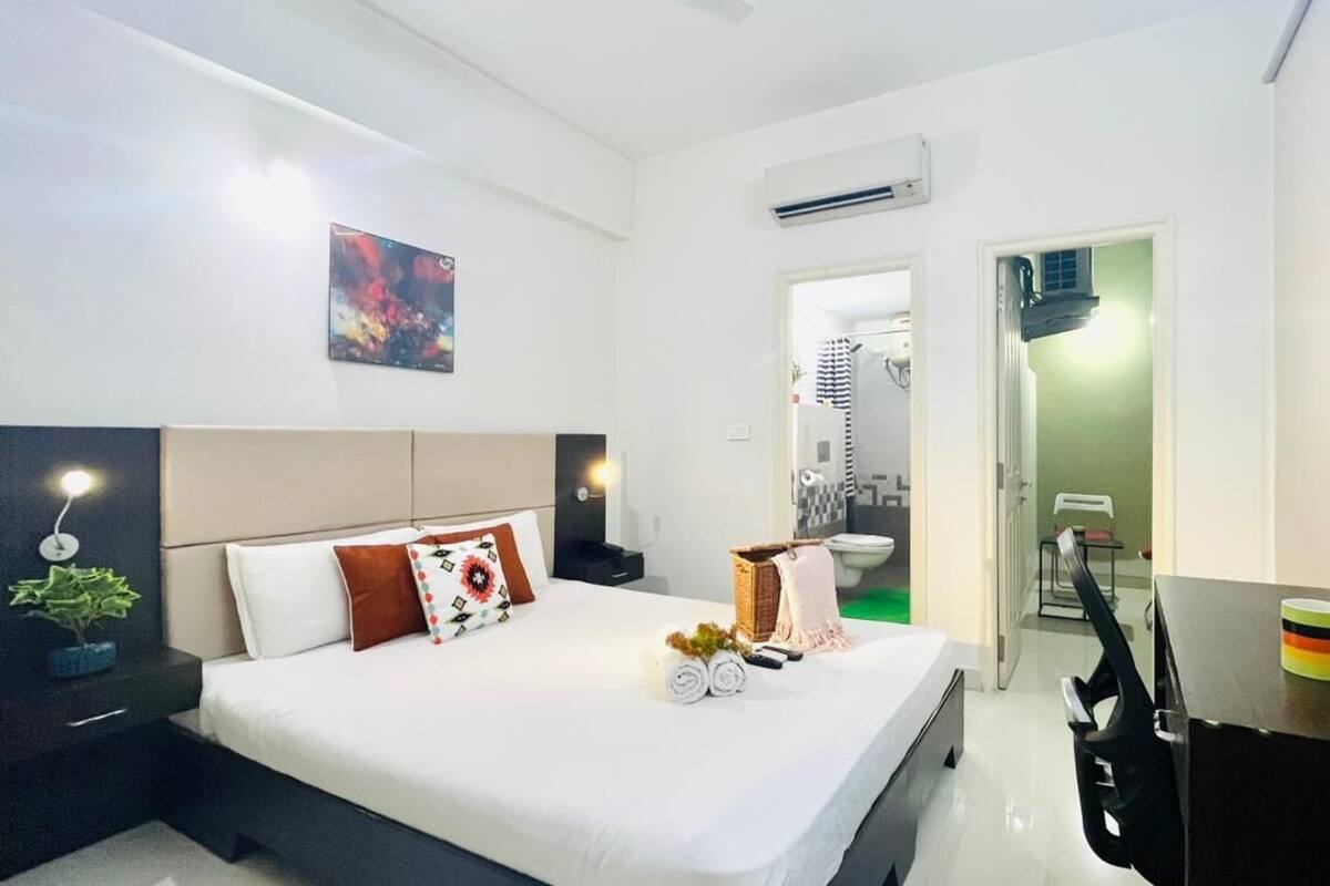 Una habitación de hotel moderna con cama, mesita de noche, televisión y un pequeño escritorio con silla.