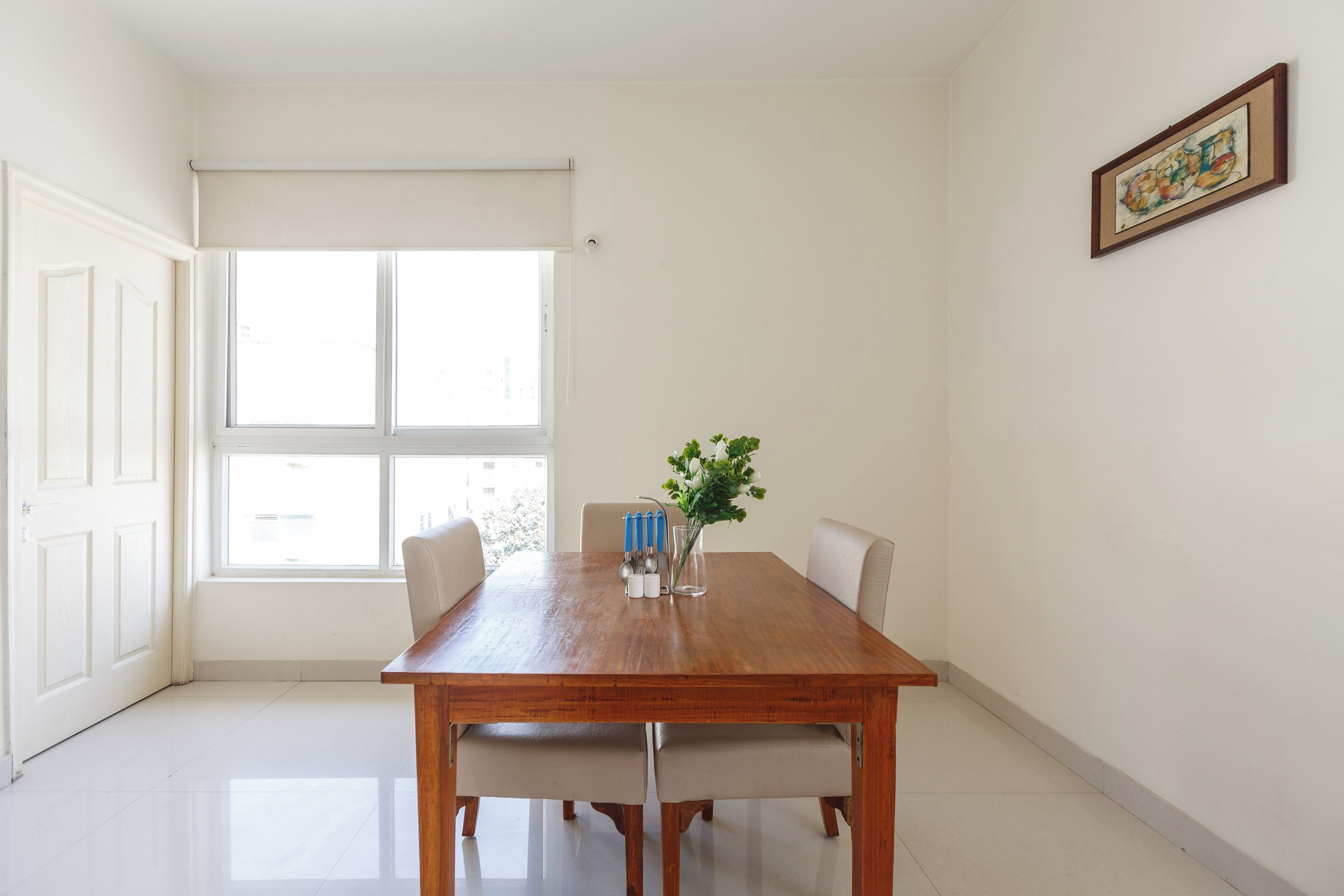 A dining area with a wooden table, four chairs, a vase with a plant, and a window with blinds.