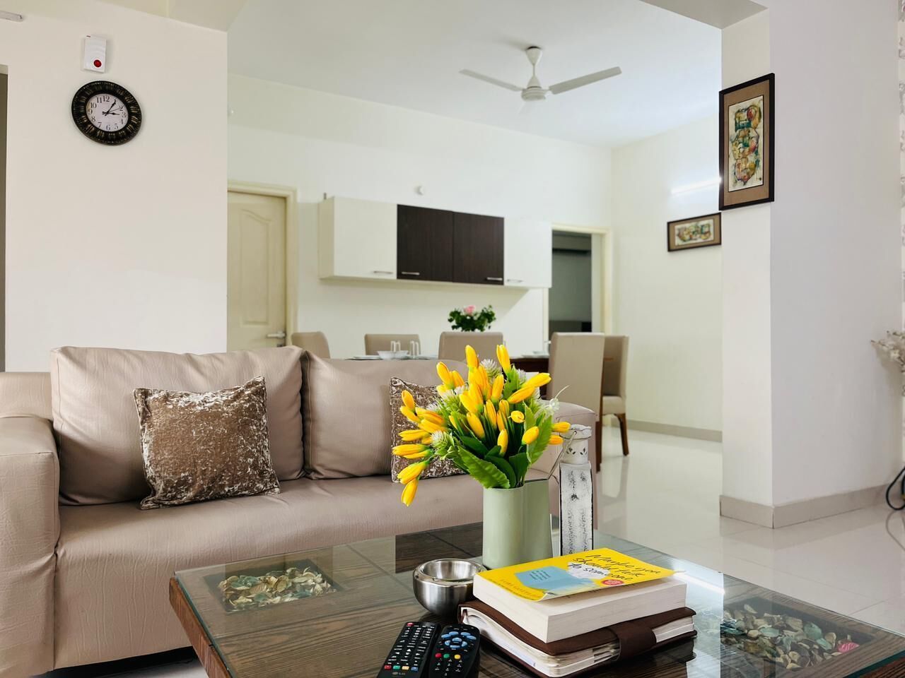 Uma sala de estar com sofá, mesa de centro com um livro e controle remoto, um vaso com flores e uma TV de tela plana ao fundo.
