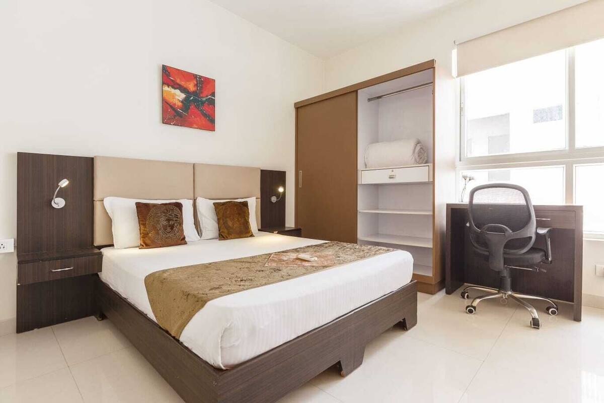 Una habitación de hotel moderna con una cama grande, un armario, un escritorio con silla y una ventana con persianas.