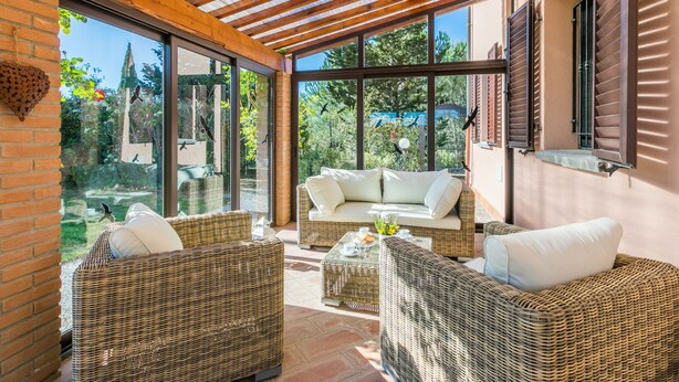 A covered outdoor seating area with wicker furniture and a glass wall providing a view of the garden. - Vacation home Lilia by Interhome (Riparbella)