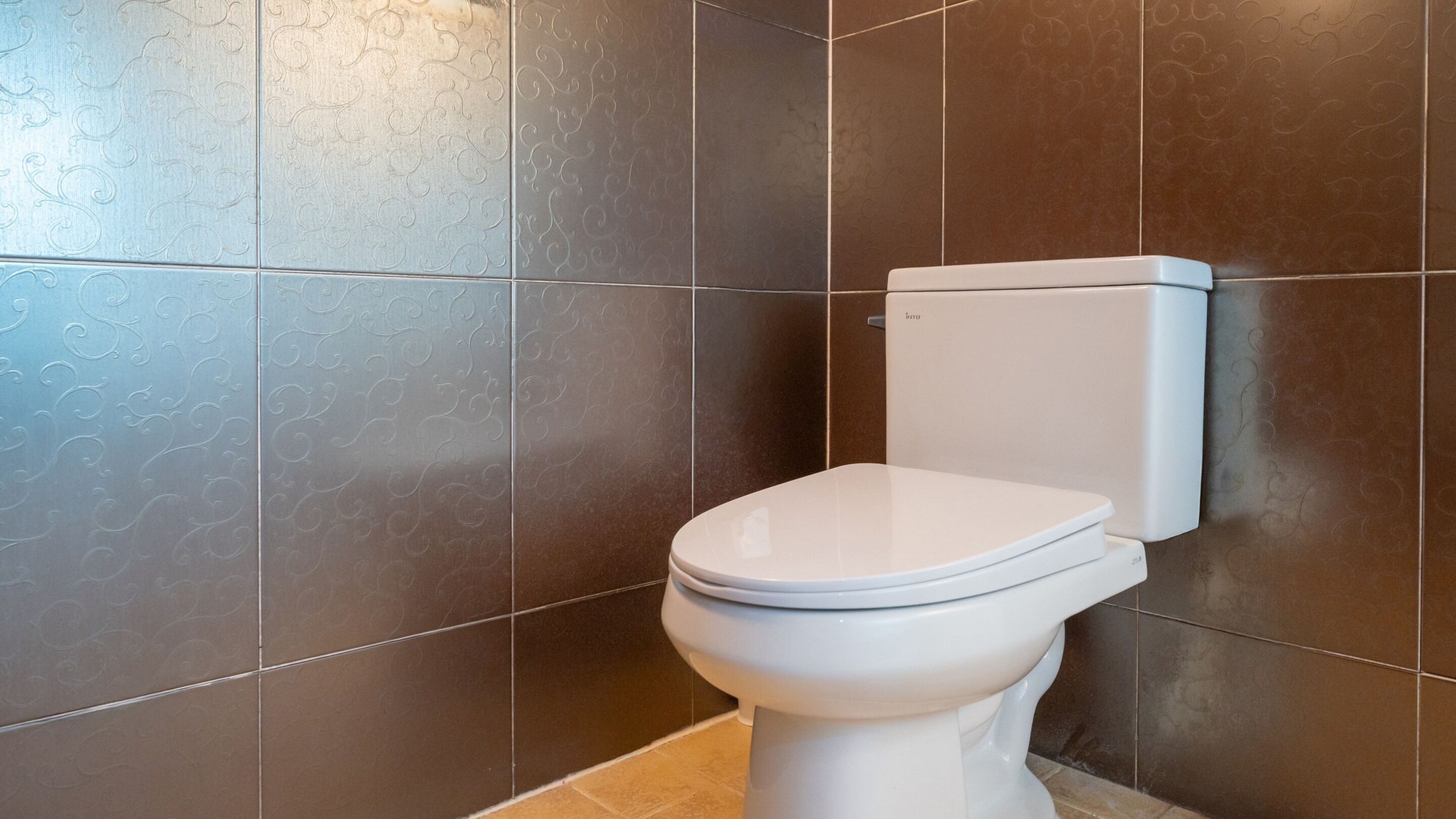 A modern bathroom with a white toilet, dark brown tiles, and a visible toilet paper holder.