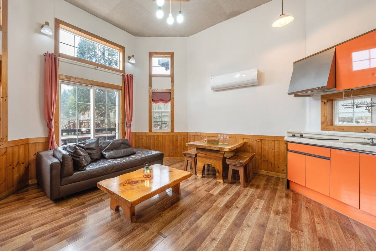 Ein gemütliches Zimmer mit Holzboden, einem Ledersofa, einem Holztisch und einer Küchenzeile mit orangefarbenen Schränken.