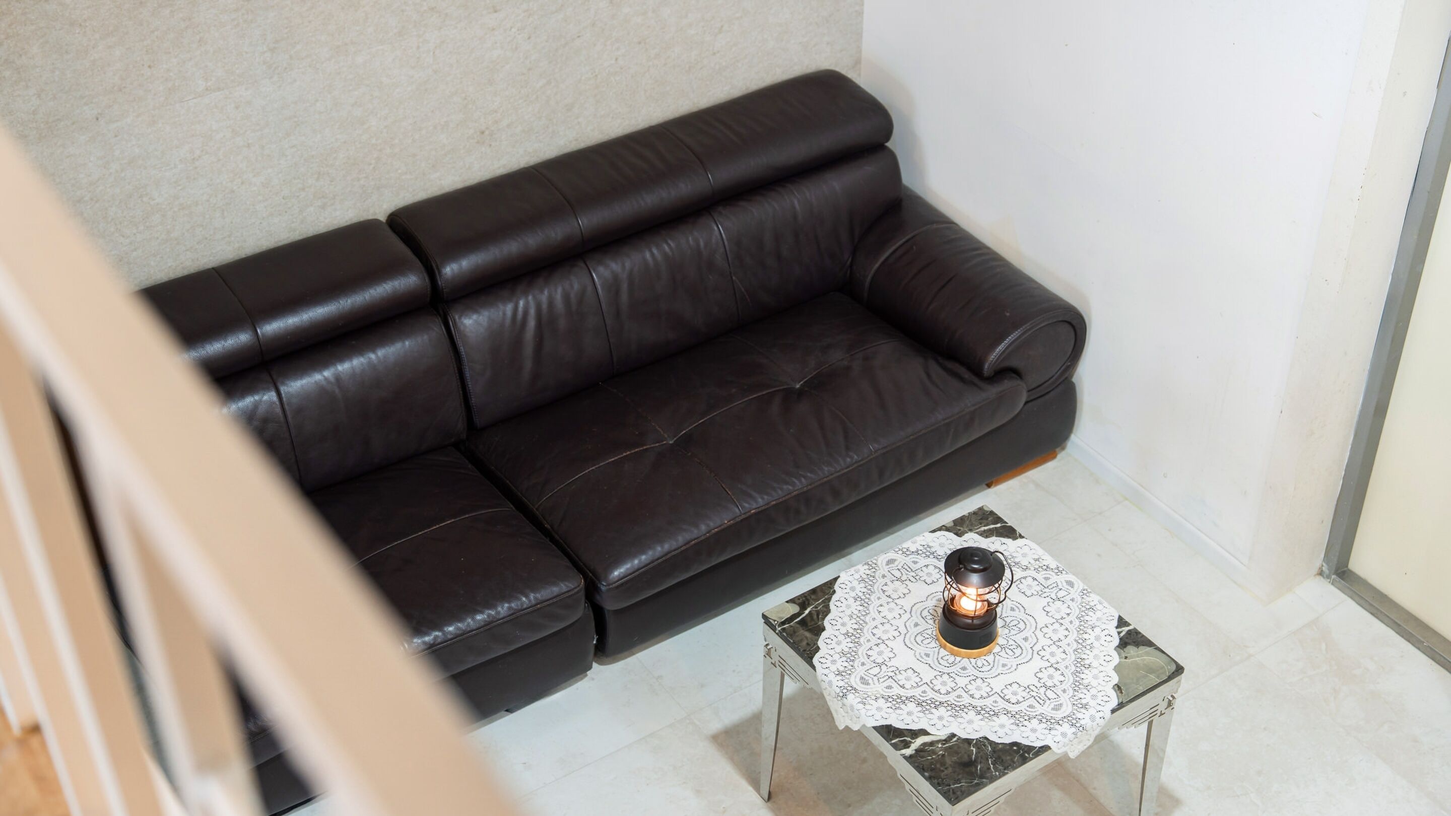 Un canapé en cuir noir adossé à un mur blanc, une petite table en verre recouverte d’une nappe en dentelle et ornée d’une bougie, et un sol carrelé.