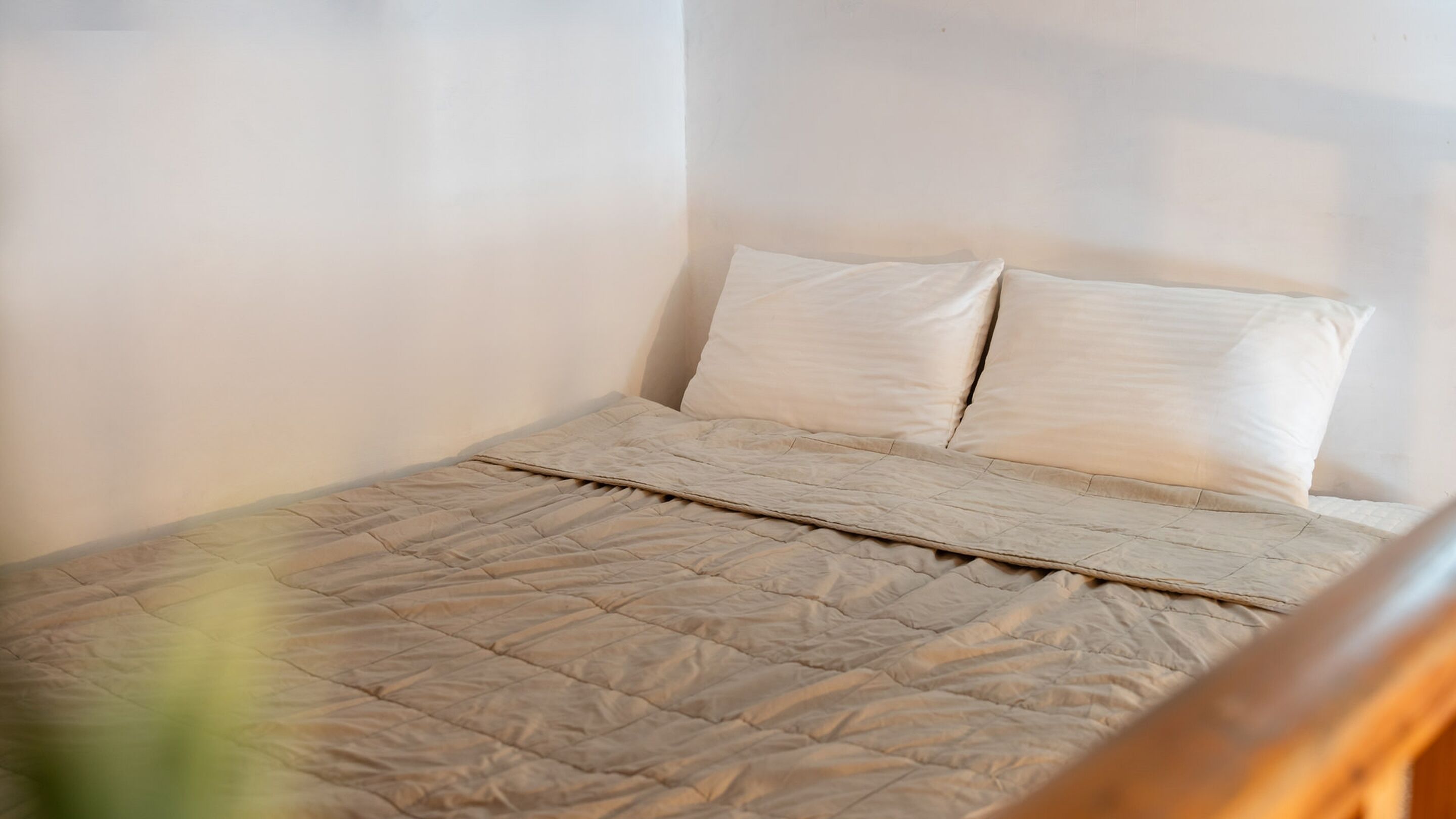 A bed with a beige quilt and two pillows, a wooden headboard, and a plain white wall.