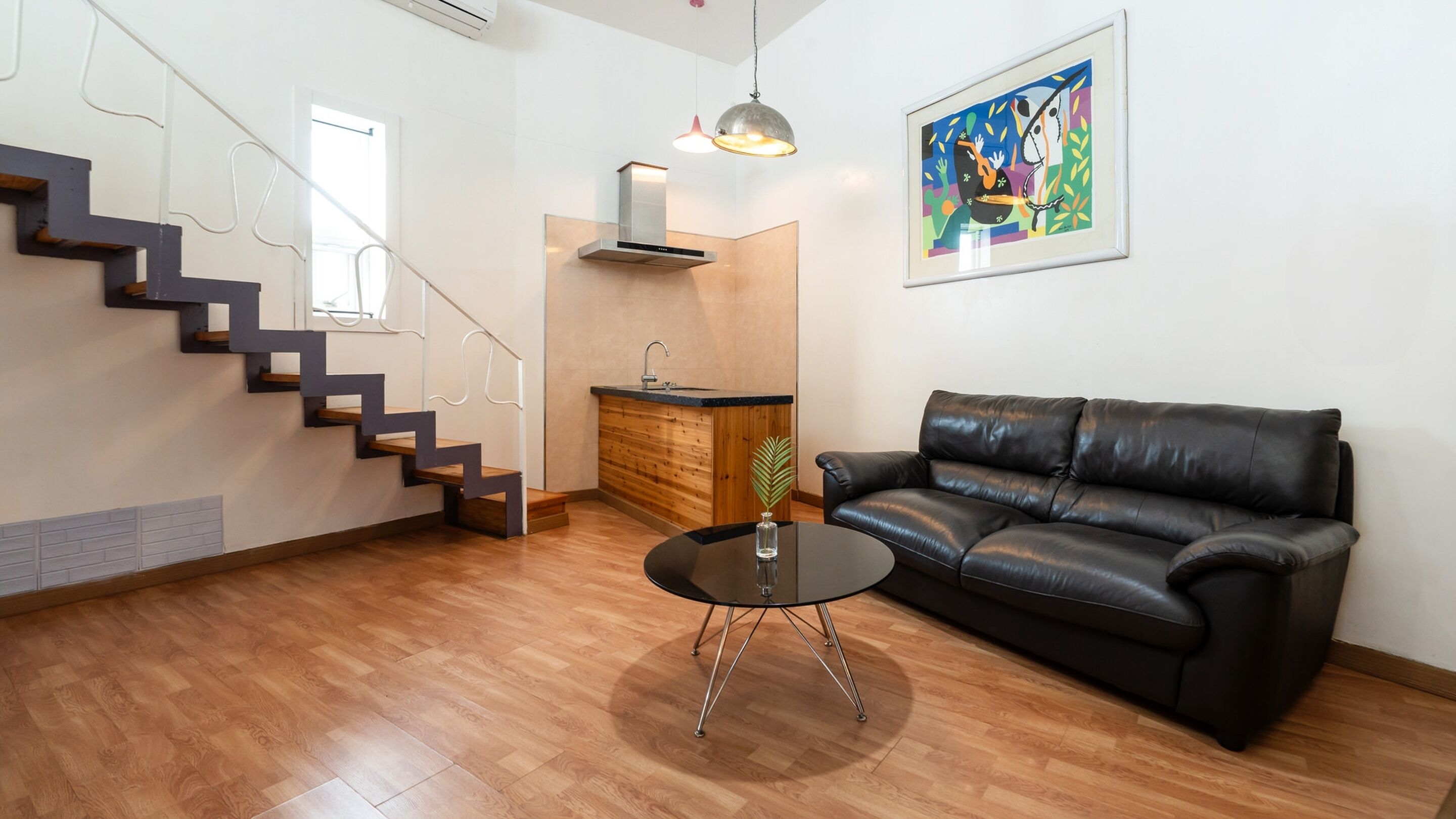 Ein modernes Wohnzimmer mit Treppe, einem schwarzen Ledersofa, einem Glastisch und einer Küchenzeile mit Holzschränken und einer Edelstahl-Spüle.