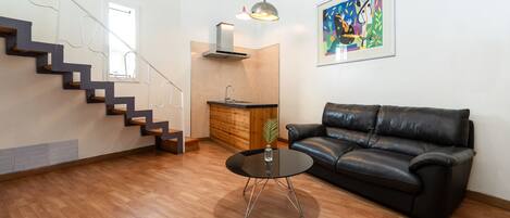 Ein modernes Wohnzimmer mit Treppe, einem schwarzen Ledersofa, einem Glastisch und einer Küchenzeile mit Holzschränken und einer Edelstahl-Spüle.