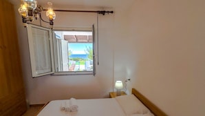 A hotel room with a bed, a nightstand with a lamp, a window with an open shutter, and a view of the outside.