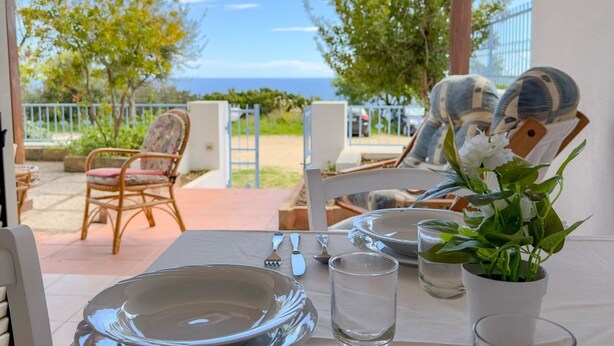 A table set for dining with a view of the sea, outdoor chairs, and a potted plant.