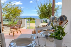Tavola imbandita per mangiare con vista sul mare, sedie da esterno e una pianta in vaso.