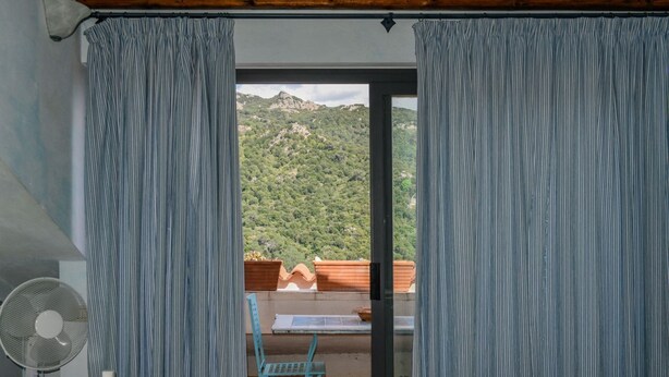 Una habitación con cama, ventilador, mesa y vistas a un paisaje montañoso a través de un ventanal.