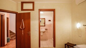 A bedroom with a bed, a bedside table, a small stool, a door leading to a bathroom with a sink and mirror. - Giardino dei Limoni Rooms 1 (Oliena)
