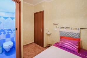 A bedroom with a bed, a bedside table, a nightstand, a wall-mounted shelf, and a door leading to a bathroom. - Giardino dei Limoni Rooms 2 (Oliena)