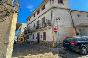 Uma rua estreita com um carro estacionado, placa de proibido estacionar e edifícios com varandas.