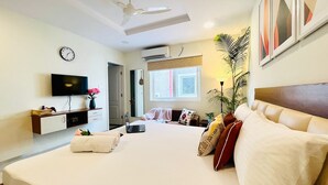 Una habitación de hotel moderna con una cama grande, un escritorio con una computadora portátil, un televisor de pantalla plana, un ventilador de techo y una ventana con persianas.