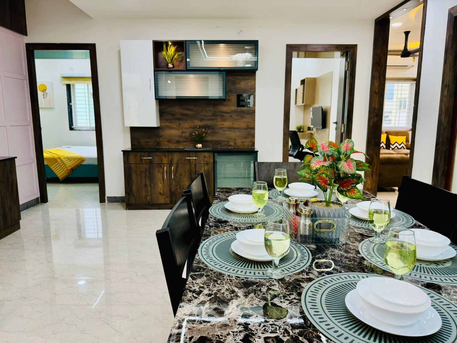 A dining area with a marble table set for four, surrounded by chairs. There is a kitchen area with wooden cabinets and a built-in oven.