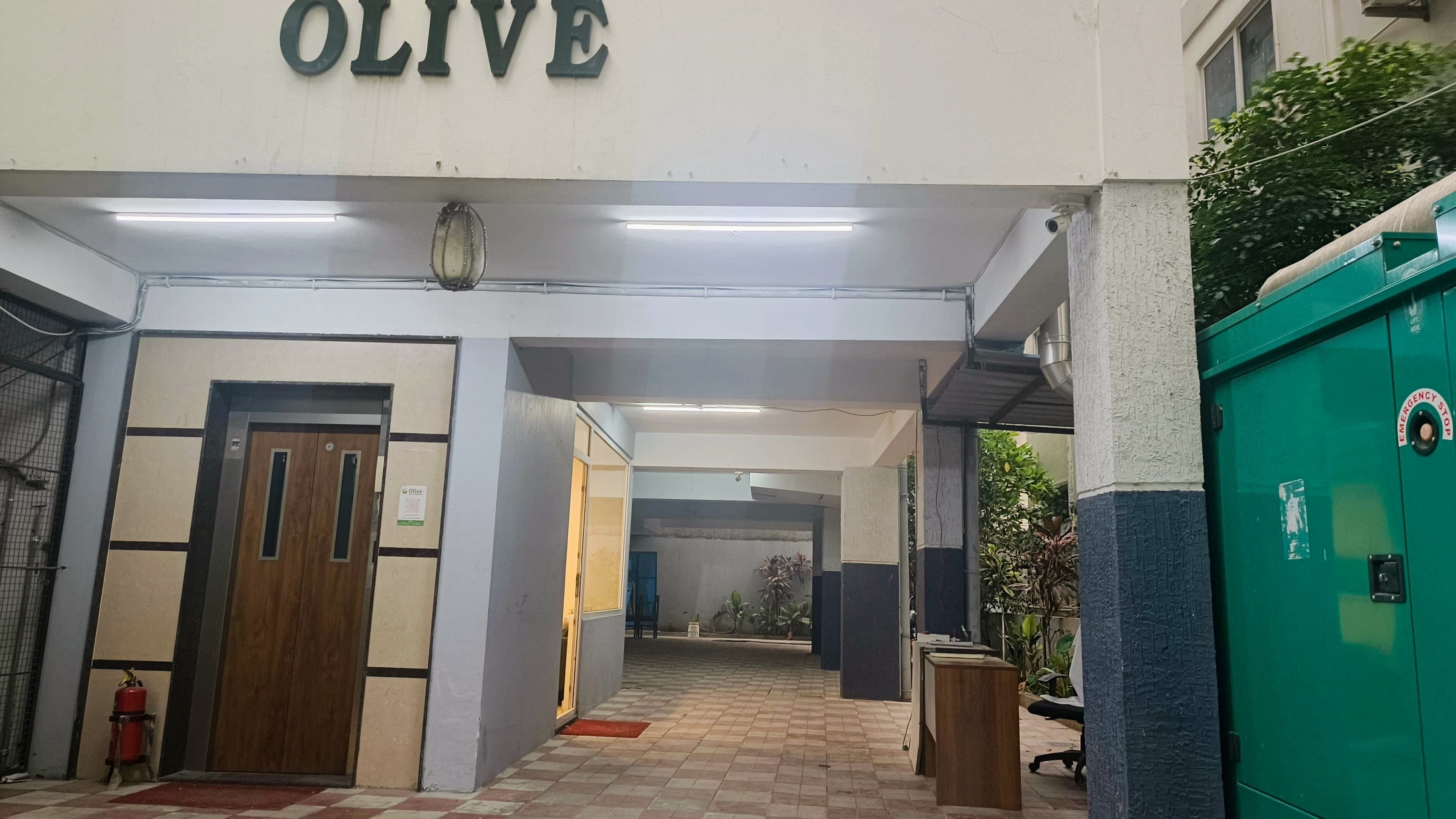 A hallway with a sign reading 'OLIVE,' a fire extinguisher, and a green emergency stop box.