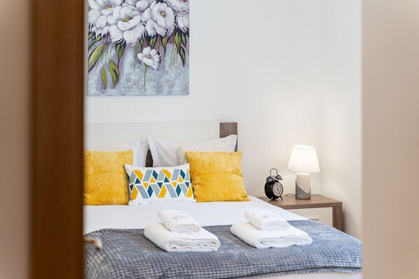 A neatly made bed with yellow and patterned pillows, a bedside table with a lamp, and a painting of flowers on the wall.