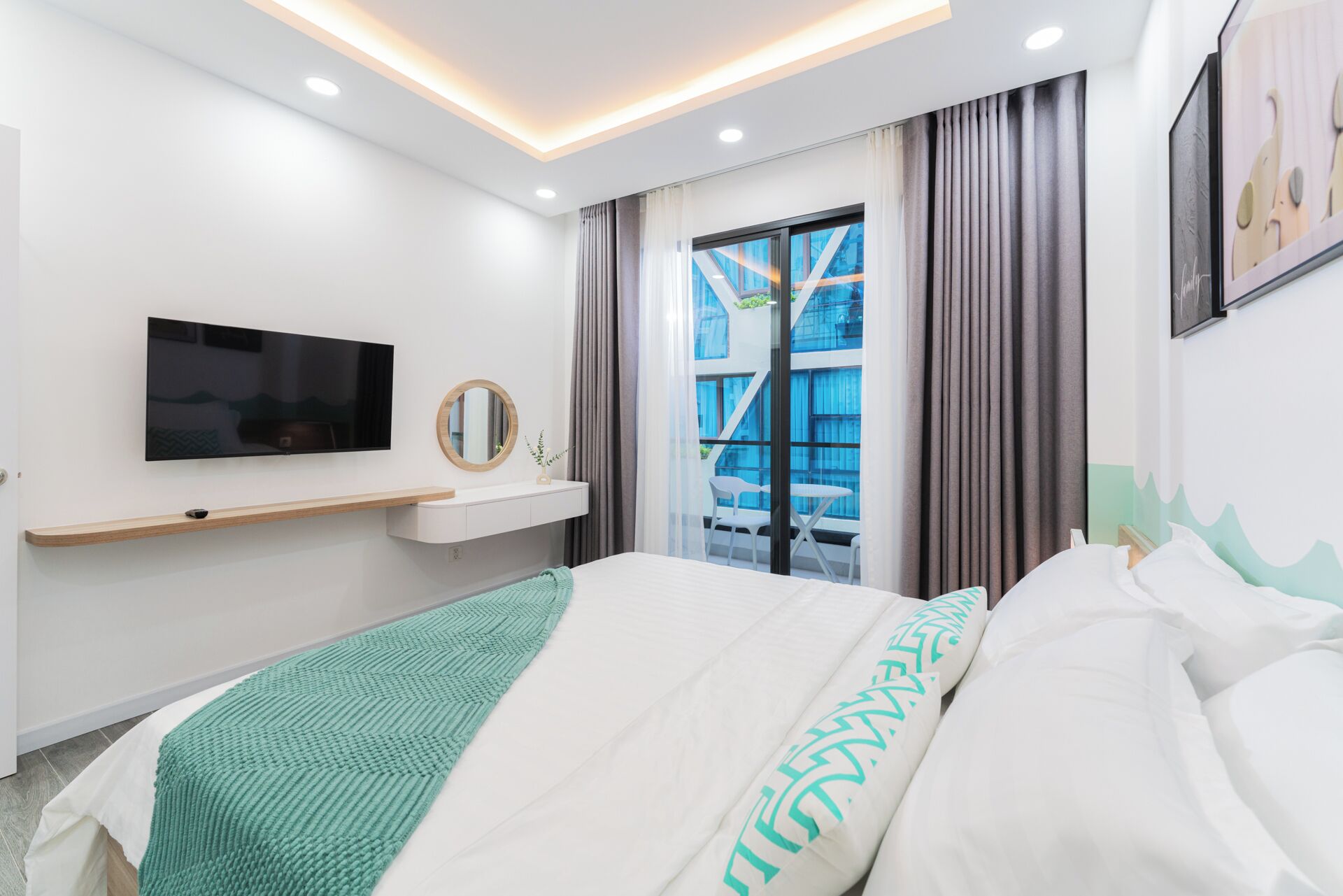 Una habitación de hotel moderna con cama, televisor montado, mesita de noche, ventana con cortinas y vistas al exterior.