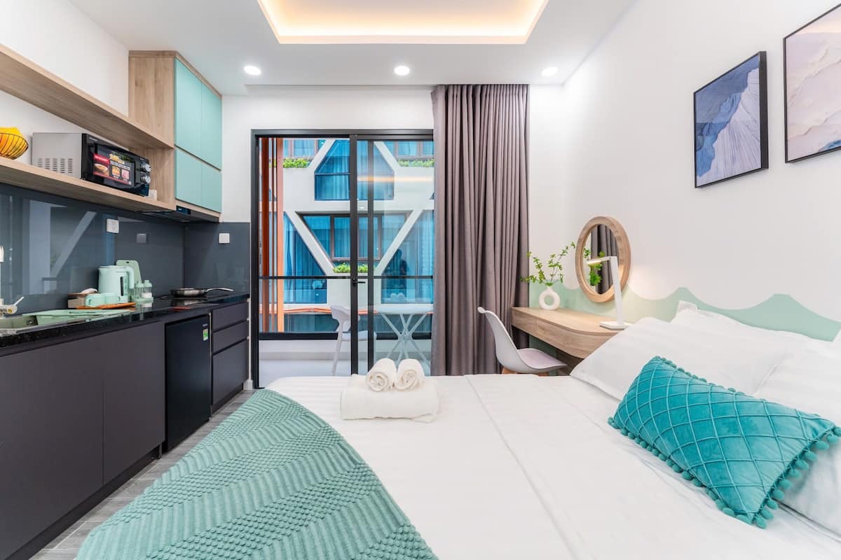 A modern hotel room with a kitchenette, a bed with teal bedding, a desk with a chair, and a balcony with a view of buildings.