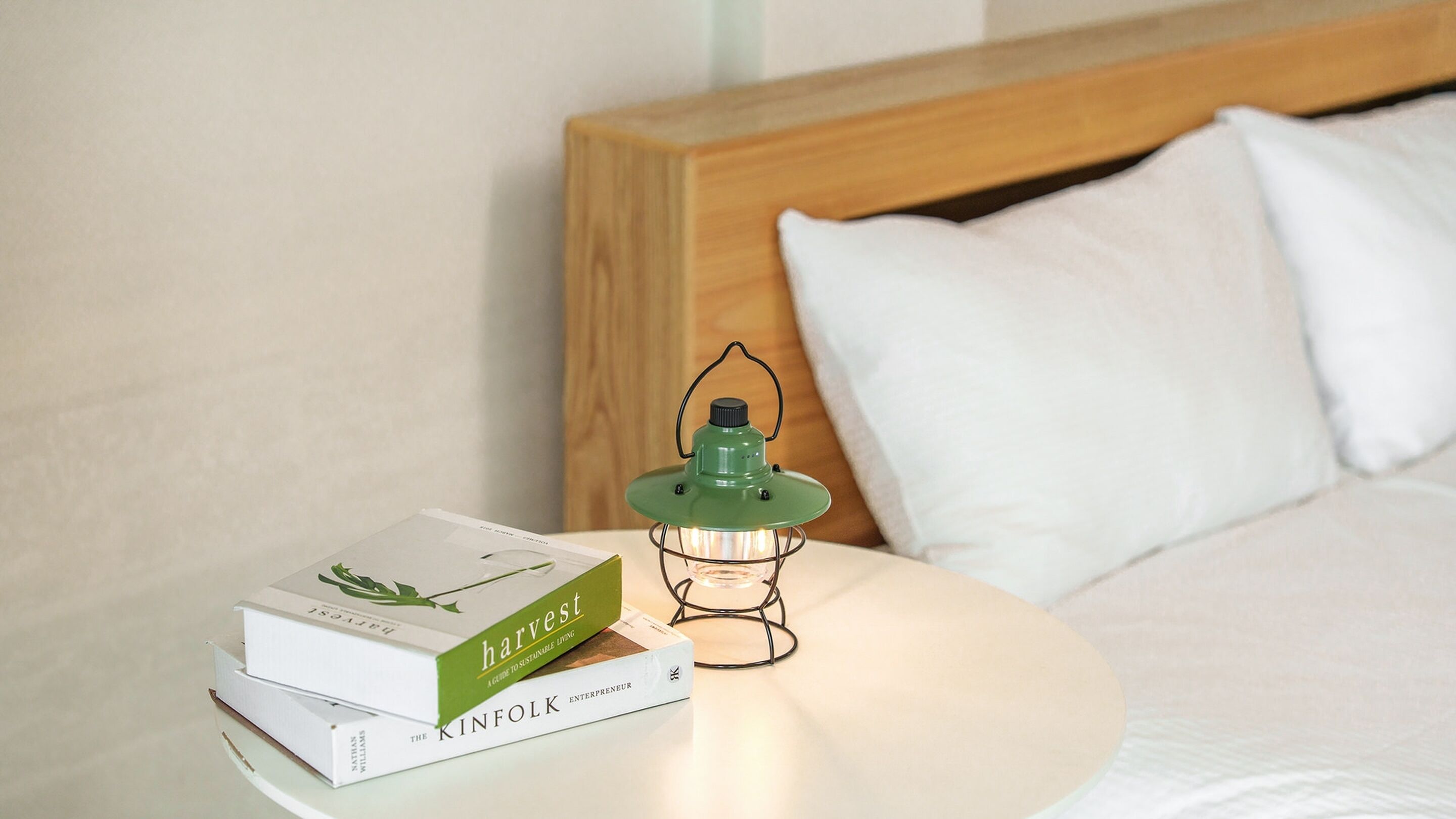 A neatly made bed with white linens, a wooden headboard, and a bedside table with a green lantern and two books titled "harvest" and "kinfolk".