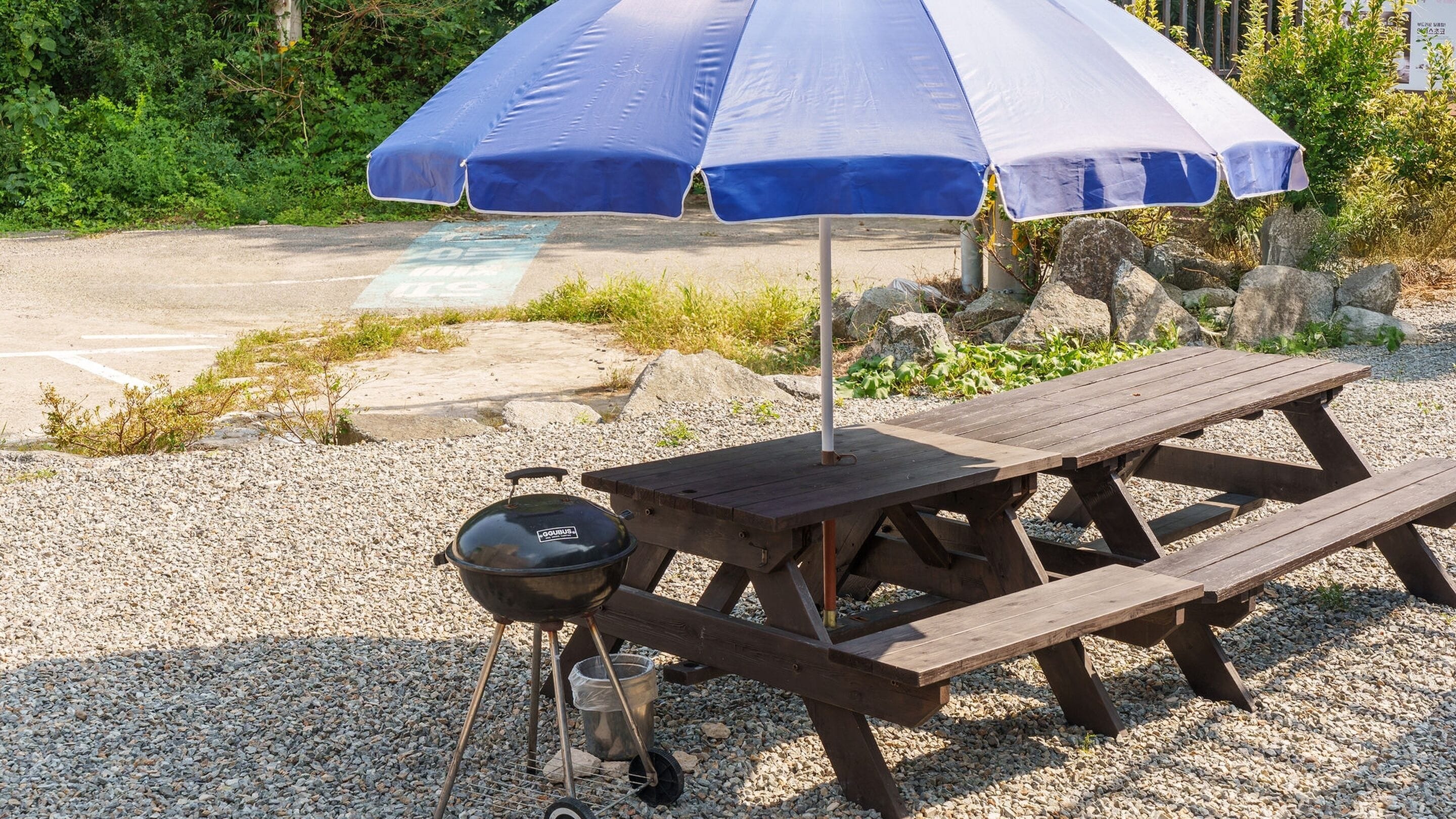 A picnic setup with a grill, a wooden table, and a blue and white umbrella.