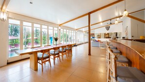 Un vaste espace dédié à la salle à manger, avec une longue table en bois et des chaises, de grandes fenêtres offrant une vue sur l’extérieur, et un coin bar avec des tabourets.