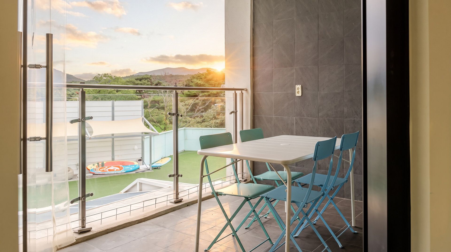 A balcony with a table and chairs, a view of a pool and greenery, and a sunset in the background.