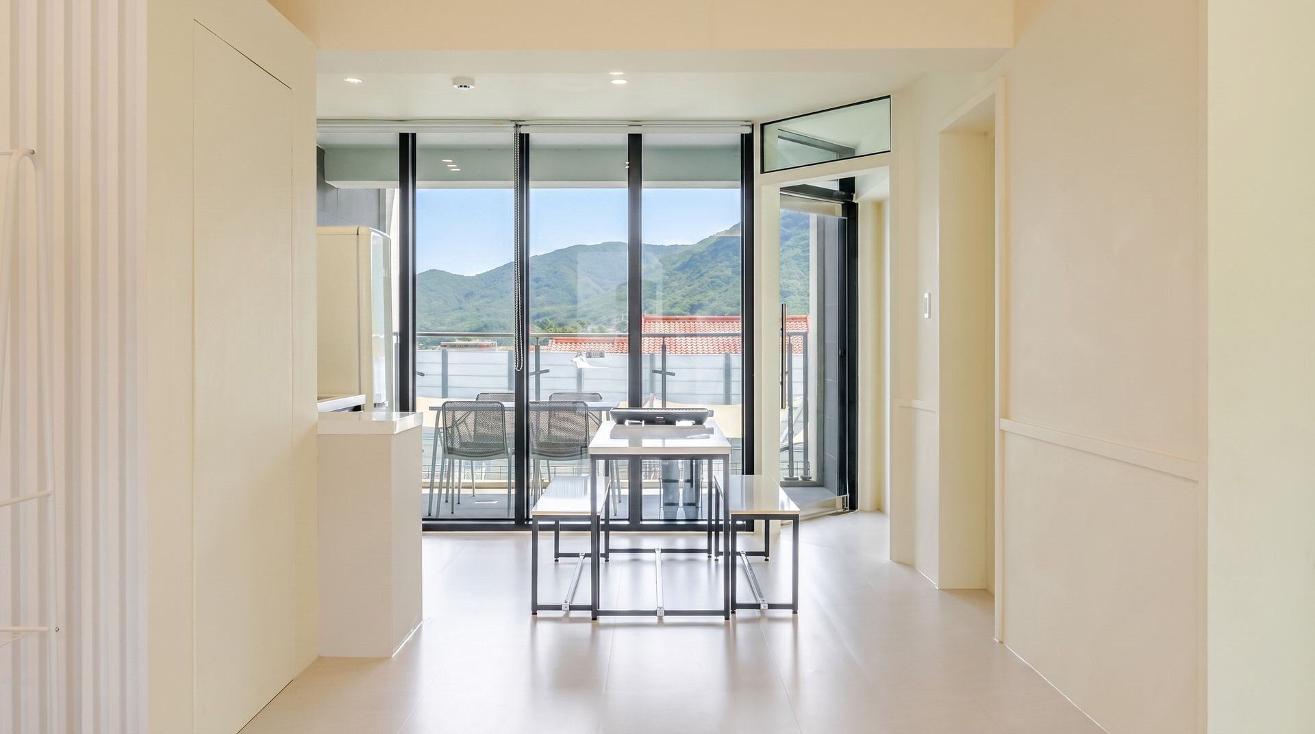 A corridor leading to a balcony with a table and chairs, a view of mountains, and a red-roofed building in the distance.