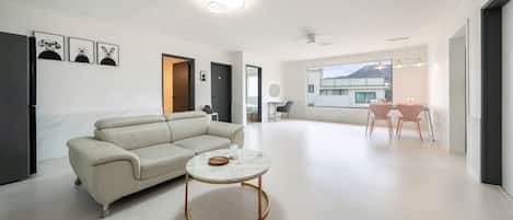 A modern living room with a sofa, a coffee table, and a dining area with chairs. There are framed pictures on the wall and a view of a building outside.