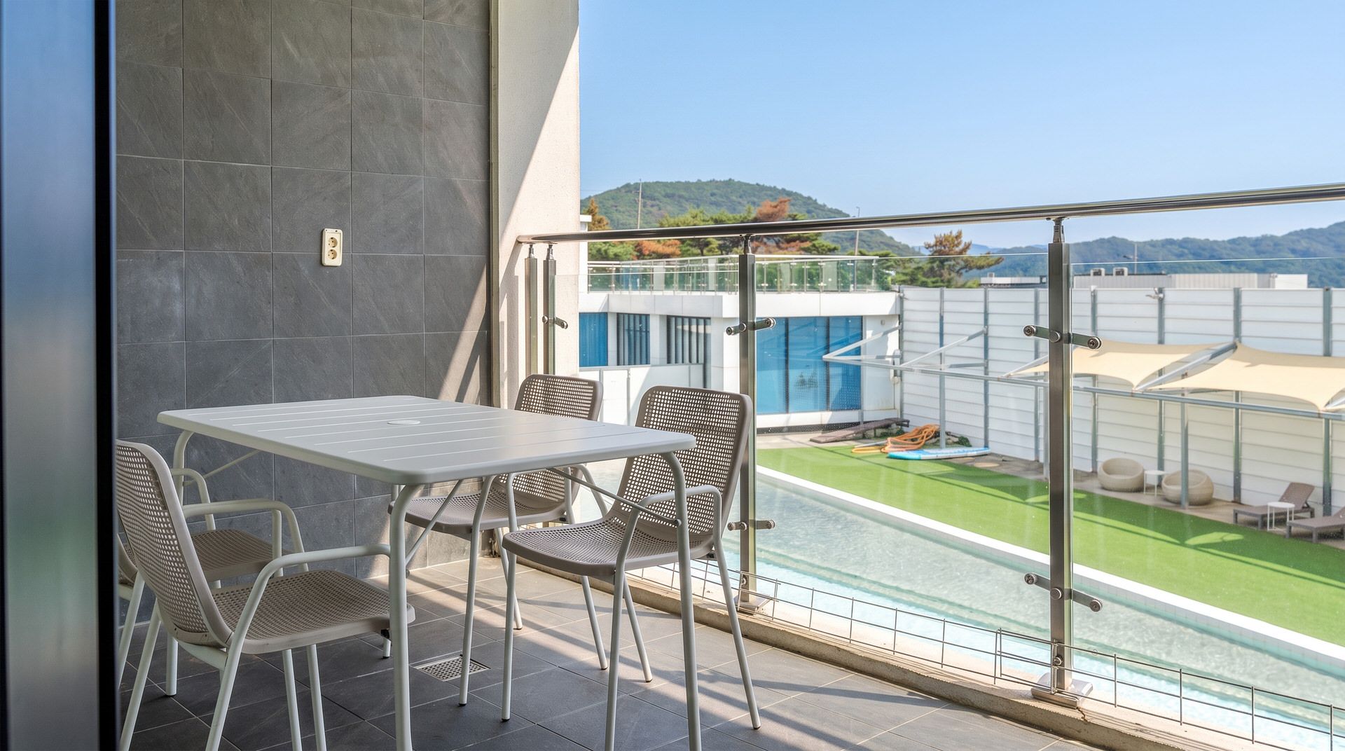 Un balcón con mesa y sillas, una piscina y vistas a un edificio y colinas.