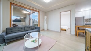 A modern apartment with a grey sofa, a glass coffee table, a pink rug, a bed, a kitchen area, and a balcony with a view.