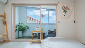 A bedroom with a large sliding glass door leading to a balcony, a bed, a small table with a picture frame, and a potted plant.
